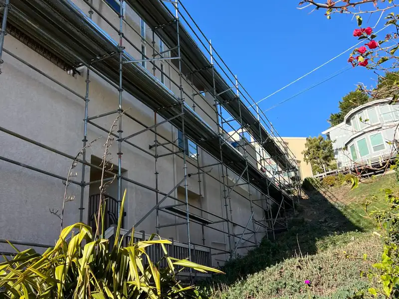 Frame scaffold along the upper floors of a white building with a flowering tree in front