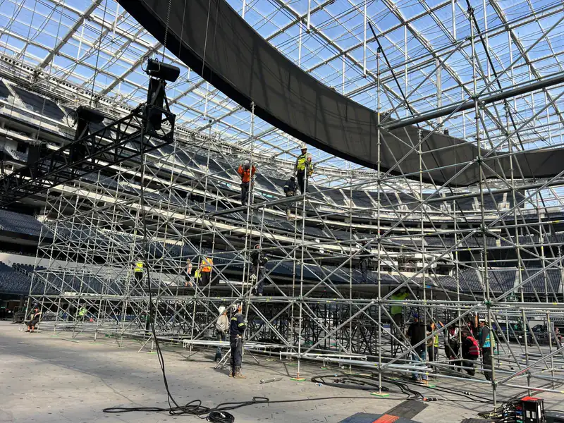 Workers building a large system scaffold under a curved glass and steel roof structure