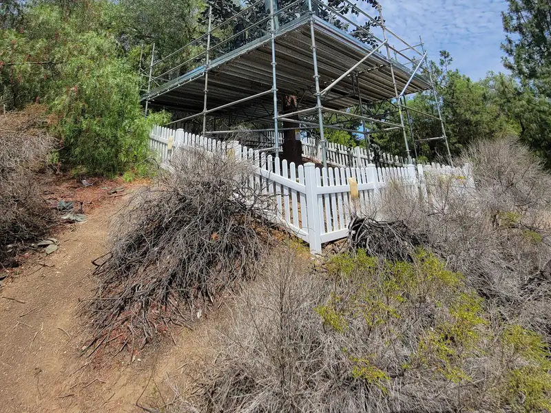 Scaffold with a covered deck built on a sloped, wooded residential lot behind a white picket fence