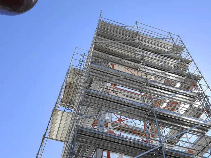 Looking up at a tall system scaffold with decked levels and orange toe boards against a blue sky